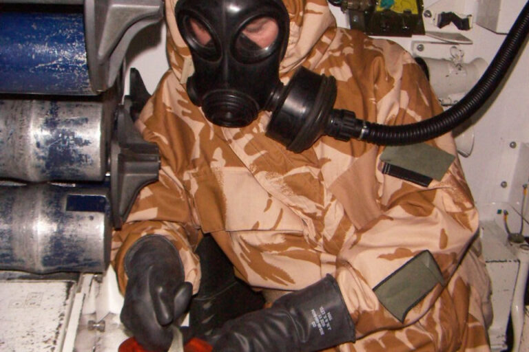 A man in a camoflauged hazmat suit wearing a gas mask and black leather gloves.